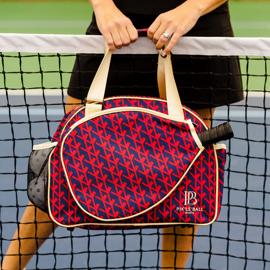 A red and navy patterned pickleball bag with cream handles is held over a tennis net by a player, with a pickleball visible in the side pocket.
