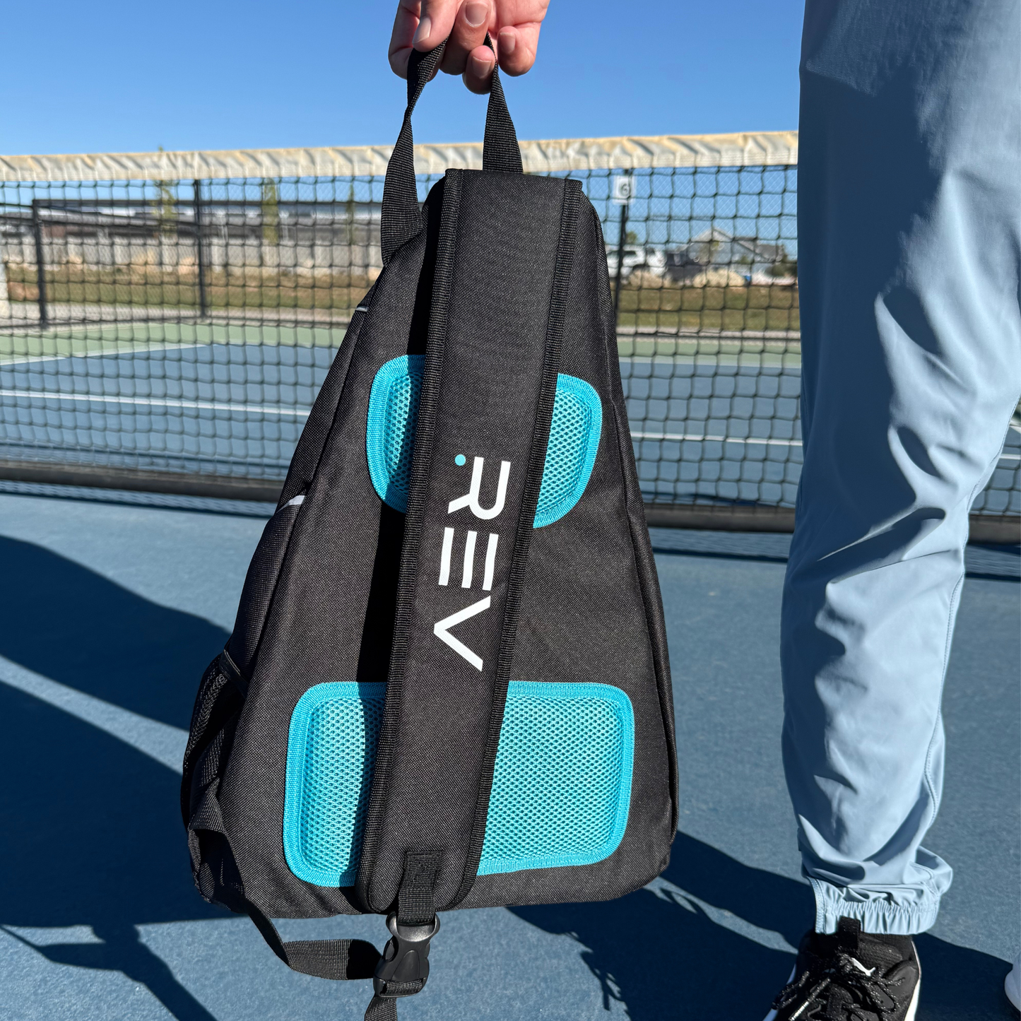 Person holding a black and blue backpack with 'REV' branding on a tennis court.