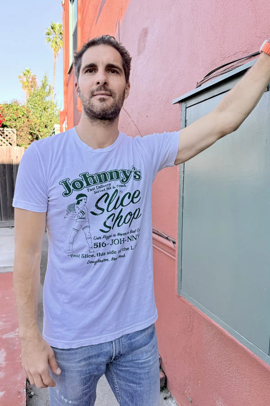Man wearing a 'Johnny's Slice Shop' t-shirt standing against a pink wall.