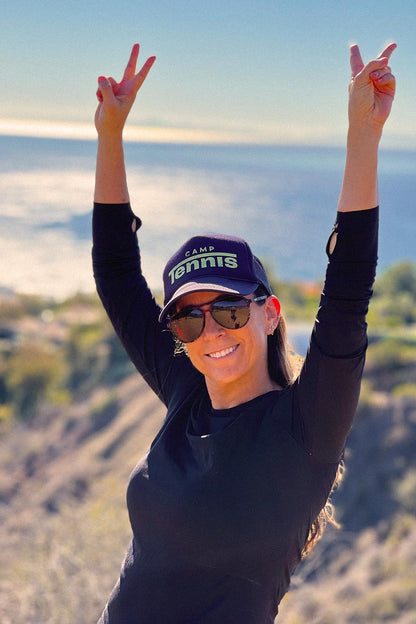 Person wearing a 'Camp Tennis' cap and sunglasses with arms raised on a beach.