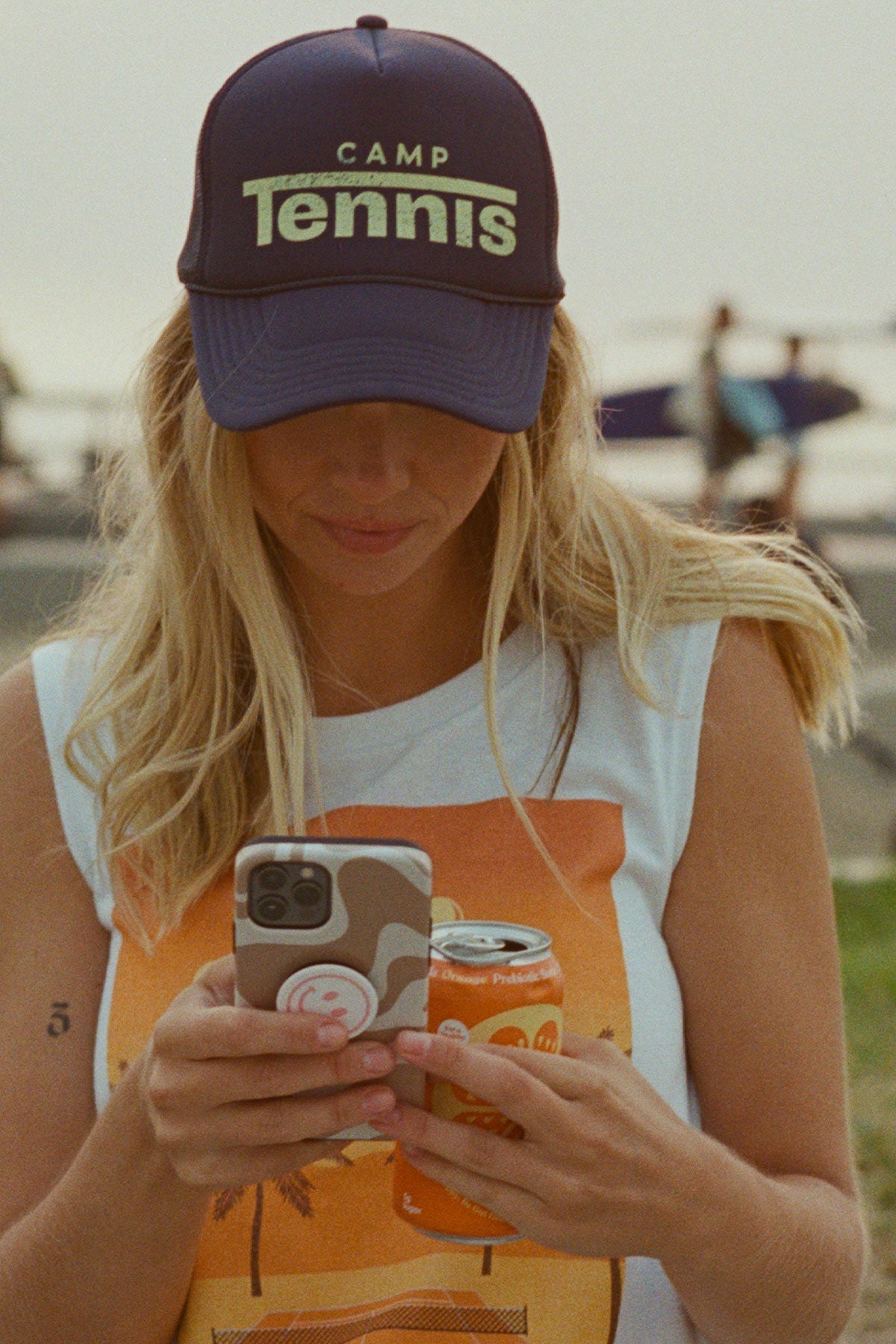 Person wearing a 'Camp Tennis' cap and holding a phone and can, with a beach background.
