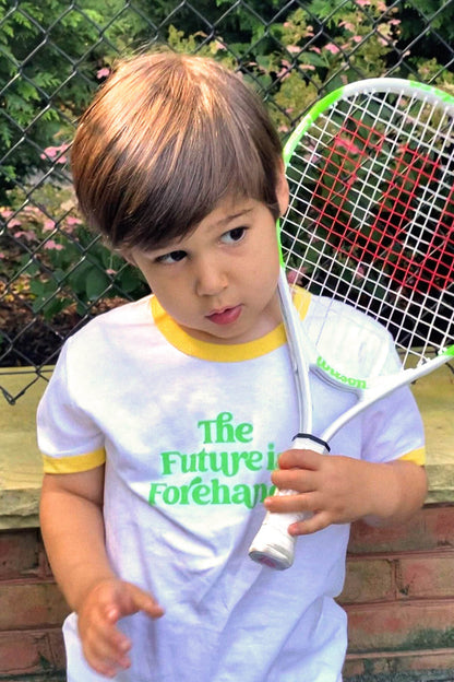 Child holding a tennis racket with 'The Future is Forehand' text on a white shirt, outdoors.