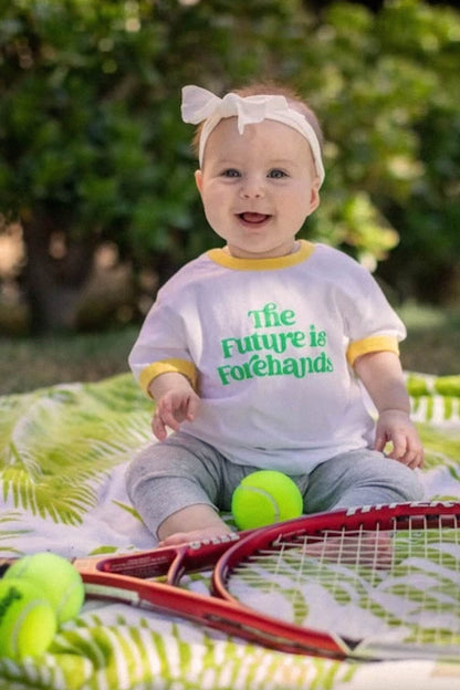 Baby sitting outdoors with tennis equipment, wearing a shirt with 'The Future is Forehands' text.