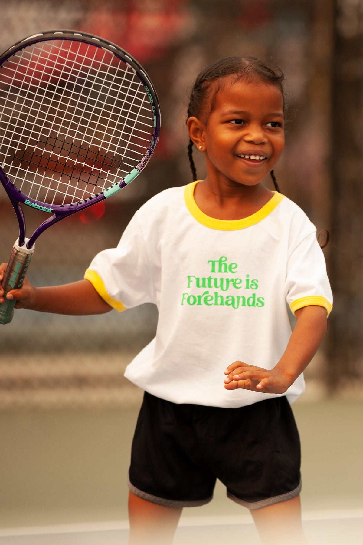 Child holding a tennis racket on a tennis court wearing a shirt with text.