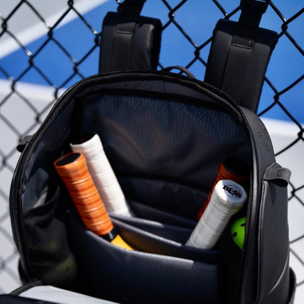 Black backpack with tennis equipment including rackets and strings on a tennis court background
