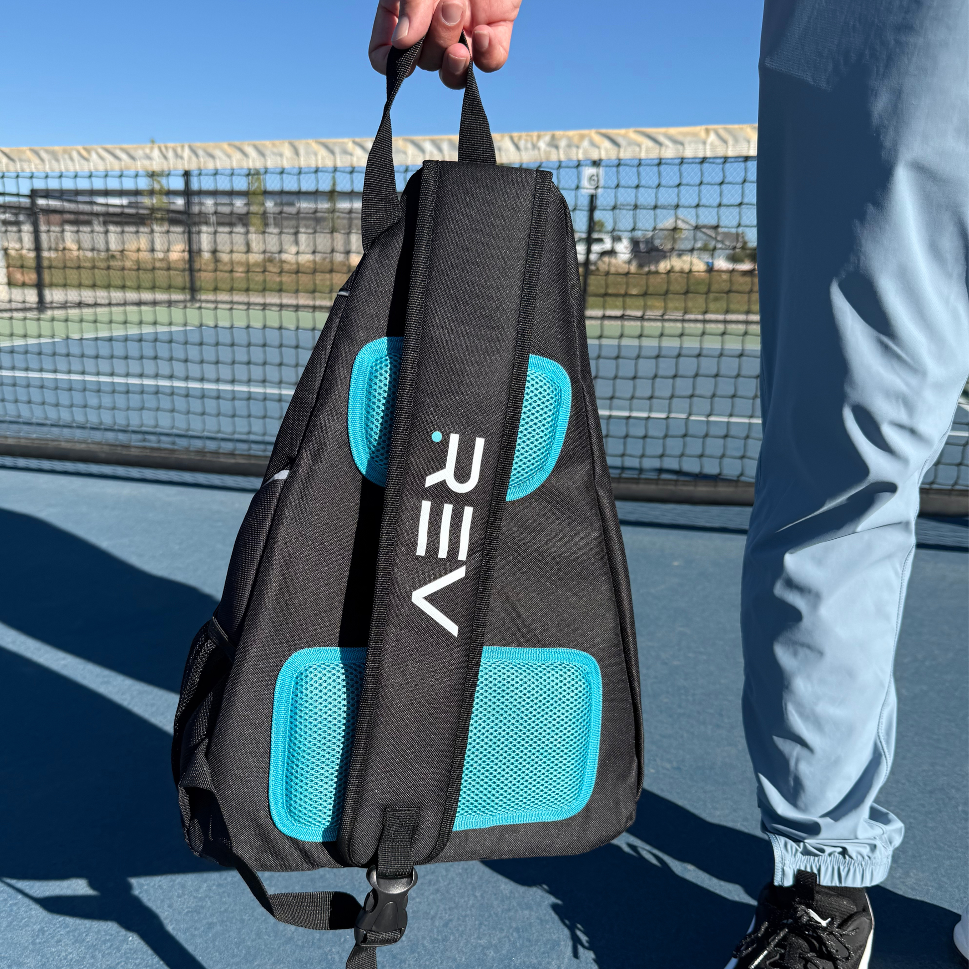 Person holding a black and blue backpack with 'REV' branding on a tennis court.