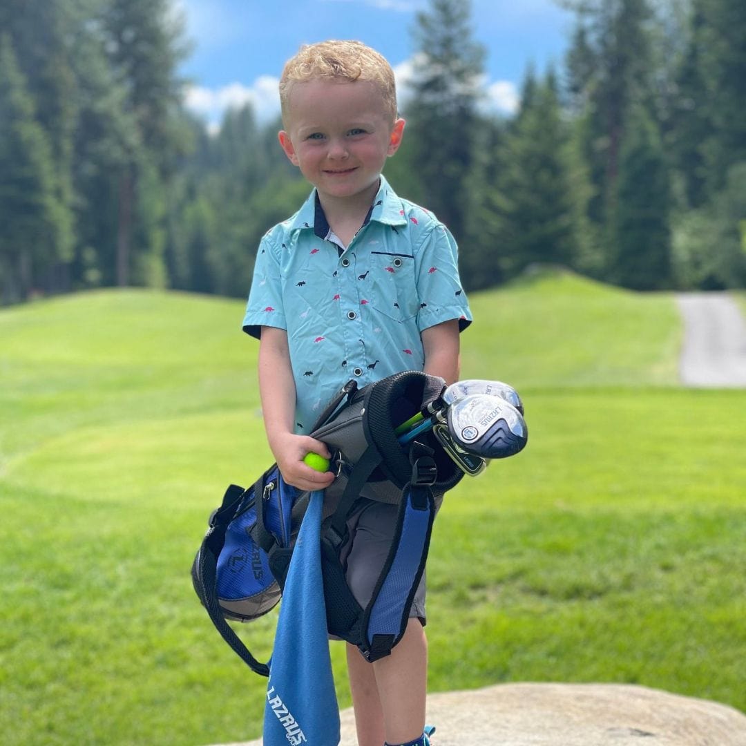 Young boy holding golf clubs and a ball on a golf course with trees in the background