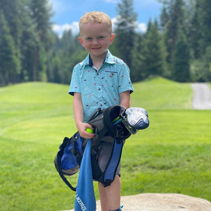 Young boy holding golf clubs and a ball on a golf course with trees in the background