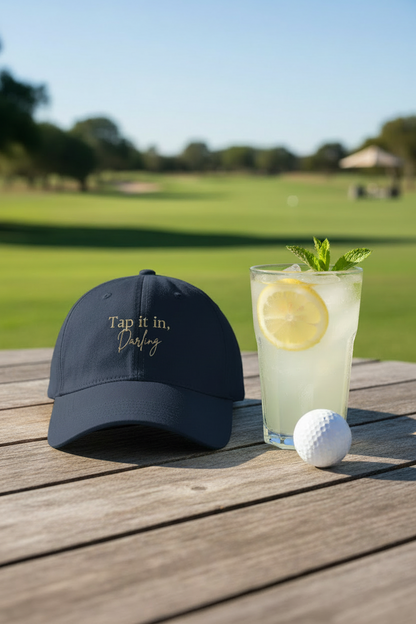 Navy cap with text, glass of lemonade with a lemon and mint, and golf ball on a wooden surface with a golf course background.