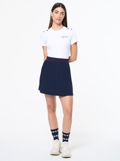 Woman wearing a white t-shirt, navy  tennis pleated skirt, and white sneakers on a white background